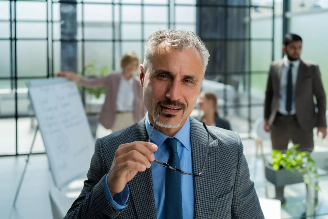 Grey-haired businessman holding his glasses in a glass-walled office, with colleagues passing behind him